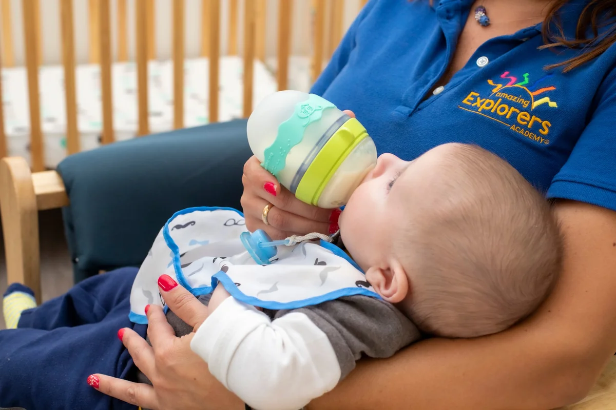Caregiver in an Amazing Explorers Academy polo bottle-feeding an infant in a rocking chair