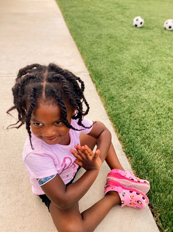 Child sitting on a sidewalk in a stretching pose beside a turf field with soccer balls